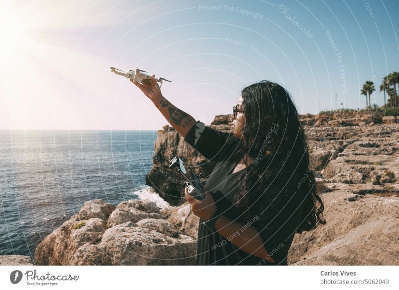 glückliche Frau mit Drohne, Blick zur Seite schwarzes Kleid und Fernbedienung, auf Klippe, am Meer sonniger Tag. technologisches Konzept Abenteuer Antenne Air