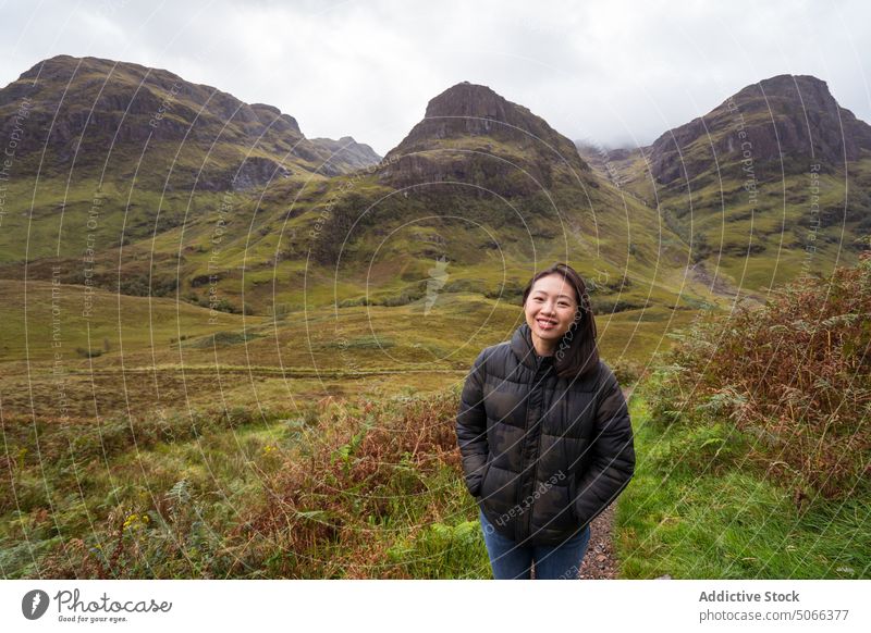Fröhlicher asiatischer Tourist vor Bergen im Tal Frau Lächeln Berge u. Gebirge Glück Oberbekleidung wolkig Natur Hochland Glen Coe Schottland Großbritannien