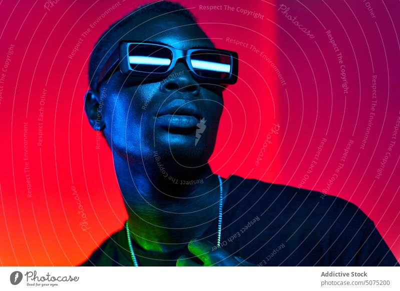 Exzentrischer schwarzer Mann mit Sonnenbrille und Kette in Neon-Beleuchtung exzentrisch neonfarbig Metaversum leuchten emotionslos flippig Stil glühen Mode