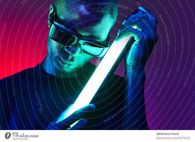 Extravaganter schwarzer Mann mit Sonnenbrille und Leuchtstofflampe in den Händen extravagant Metaversum reflektieren neonfarbig Lampe fluoreszierend glühen