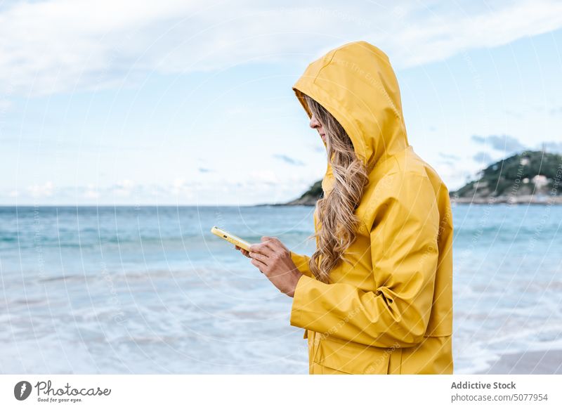 Frau, die am Strand auf ihrem Smartphone surft Browsen Meeresufer Küste Regenmantel MEER Surfen Internet Mobile Telefon Textnachricht gelb Spanien Anschluss SMS