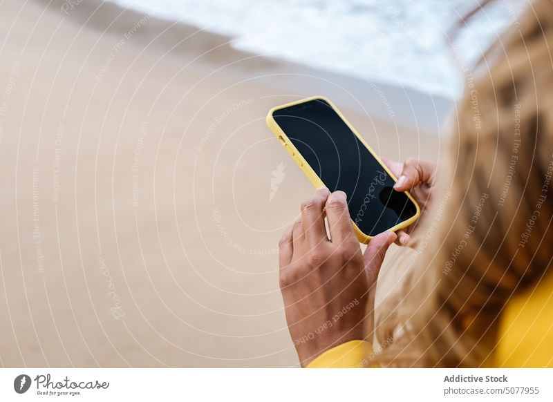 Anonyme Frau, die am Strand auf ihrem Smartphone surft Browsen Meeresufer Küste Regenmantel MEER Surfen Internet Mobile Telefon Textnachricht gelb Spanien