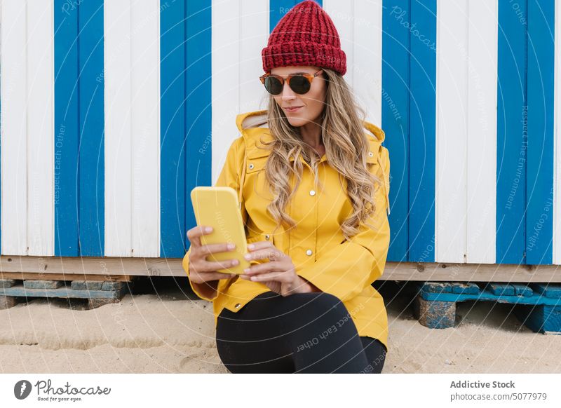 Modische Frau mit Sonnenbrille und Regenmantel macht ein Selfie am Strand Smartphone Stil Meeresufer farbenfroh Gedächtnis reisen blättern trendy blond Mobile