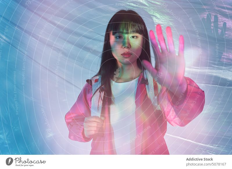 Asiatische Frau zeigt Stopp-Geste in futuristischem Licht Chinesisch neonfarbig emotionslos Handfläche stoppen glühen modern Rucksack leuchtend im Freien