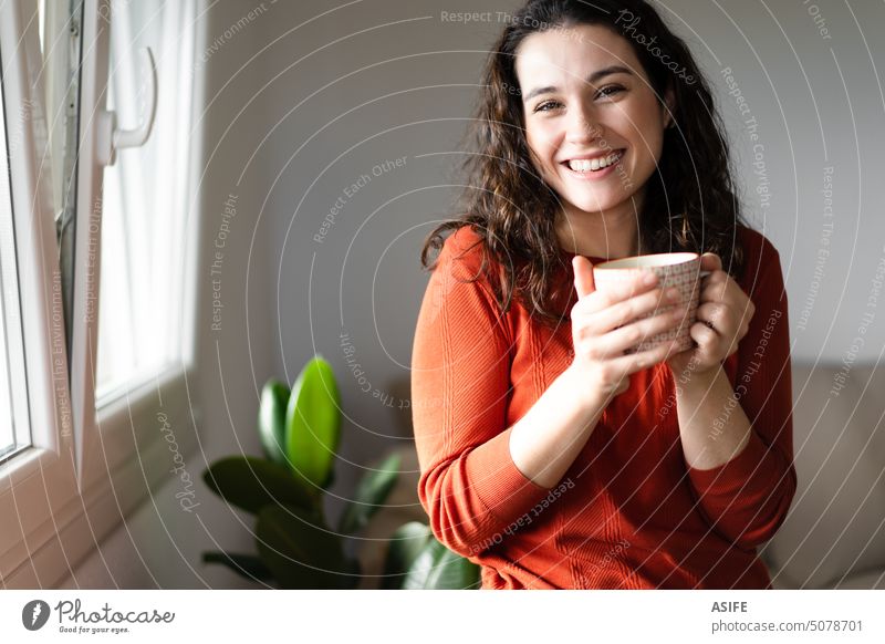 Glückliche junge schöne Frau, die einen Kaffee oder Tee zu Hause trinkt heimwärts Lächeln Lachen in die Kamera schauen zahnfarben Beteiligung Tasse trinken