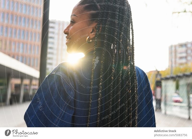 Schwarze Frau mit Afrozöpfen steht auf der Straße Großstadt Quadrat Gebäude Afro-Zöpfe urban Kälte Sonnenschein tagsüber Afroamerikaner schwarz ethnisch jung
