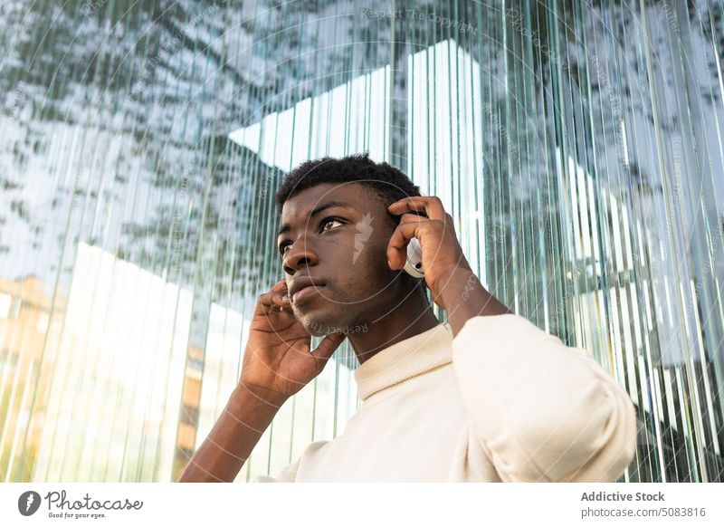Schwarzer Mann mit Kopfhörern in der Nähe eines modernen Gebäudes zuhören Musik benutzend Drahtlos ernst cool nachdenklich Apparatur Großstadt männlich Gerät