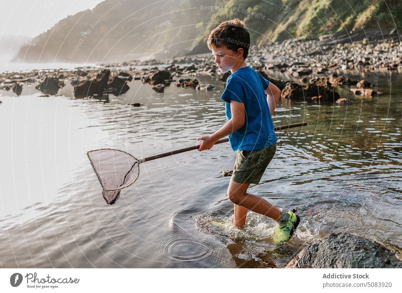 Junge mit Fischschaufel am Felsenufer Netz Baggerlöffel Ufer Küste Sommer Fischen Kind Kindheit Fischer seicht Aktivität sorgenfrei Hobby Sonnenlicht Stein