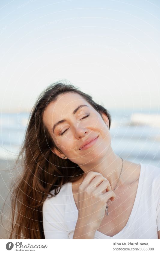 junge erwachsene kaukasische Frau lächelnd friedlich am Strand in der Nähe von Meer mit geschlossenen Augen. Weibliche candid Porträt der entspannten Person. Kaukasische Frau mit braunem Haar.