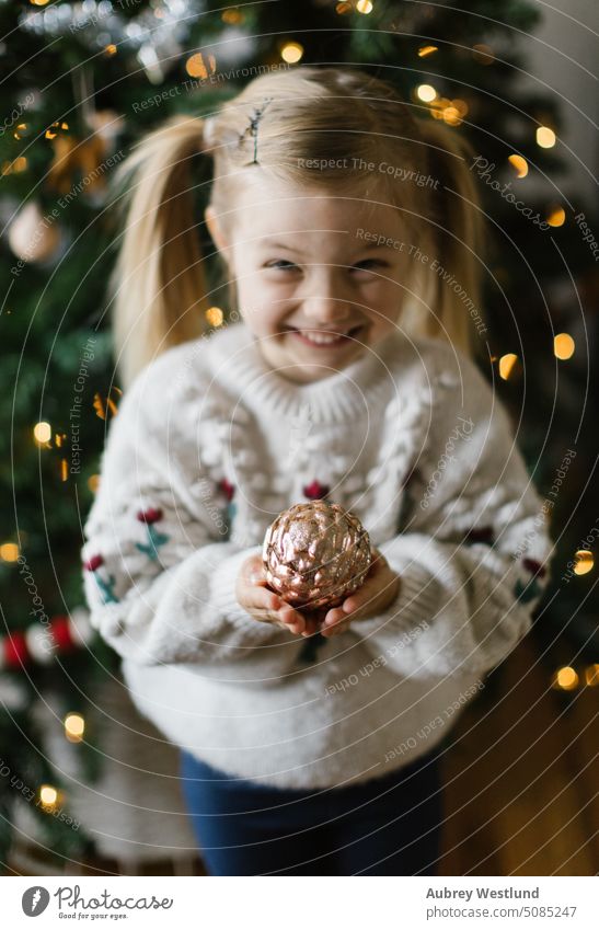 Kleines Mädchen hält goldenes Ornament vor einem Weihnachtsbaum Weihnachtsmann Hintergrund blond hell Feier Kind Kindheit Weihnachten niedlich Dezember