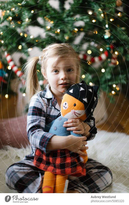 Kleinkind im karierten Schlafanzug, das mit seinem Stofftier vor dem Weihnachtsbaum kuschelt Weihnachtsmann Hintergrund blond hell Feier Kind Kindheit