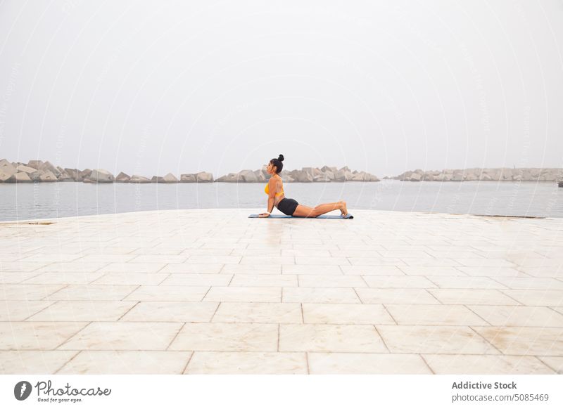 Sportliche Frau in Cobra-Pose auf Matte am Meer mit Steinen Yoga Kobra-Pose bhujangasana Asana Training Übung Achtsamkeit Konzentration Ausdauer Bestimmen Sie