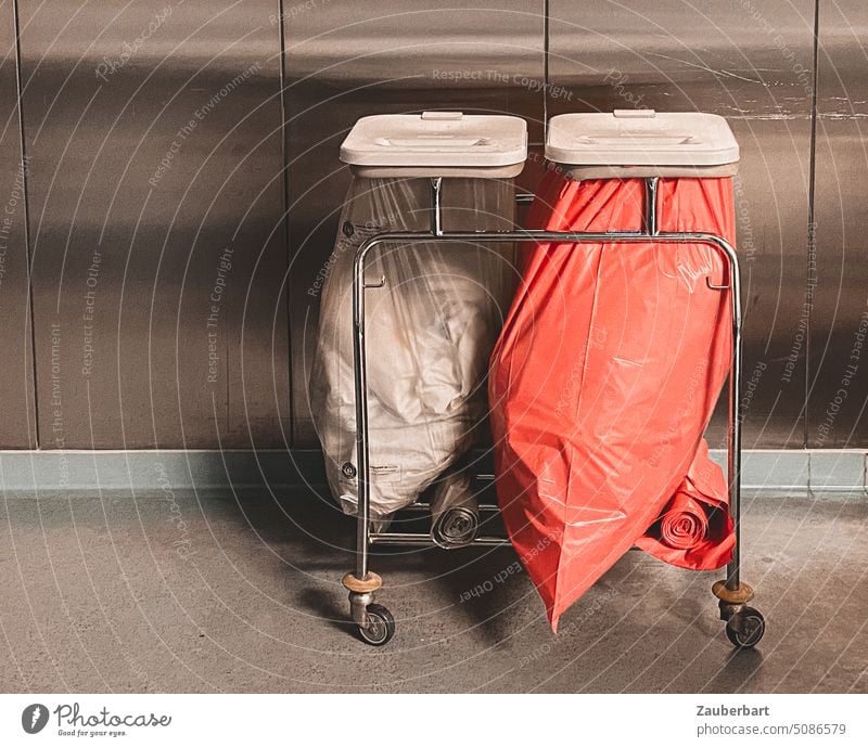 Abfallsammler mit zwei Mülltüten, gefüllt mit Verbandsmaterial, vor einem Wandschrank aus Metall in einem Krankenhaus Rollwagen rot transparent Metallwand