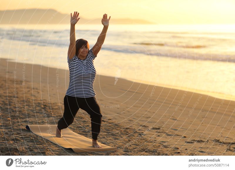 Reife Frau kümmert sich um den Körper und macht Yoga am Meer außerhalb auf Sonnenaufgang. Weibliche gekleidet den Sport tragen und tun Sportübungen in Sandstrand. Meditation, Yoga und Entspannung Konzept.