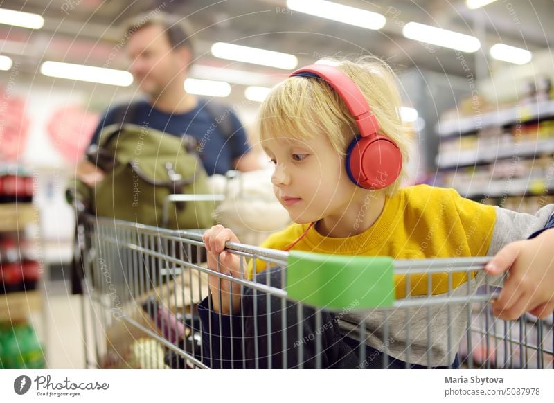 Niedlicher Junge im Vorschulalter mit Kopfhörer und Player sitzt in einem Einkaufswagen in einem Lebensmittelgeschäft oder Supermarkt. Ein Kind hört Musik oder ein Hörbuch, während seine Eltern Lebensmittel einkaufen.