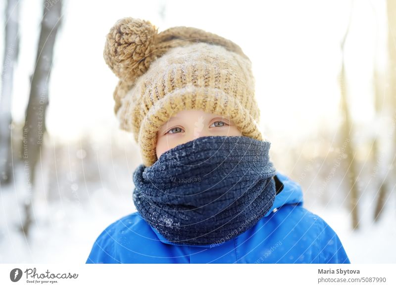 Porträt des kleinen Jungen in blauer Winterkleidung Spaziergänge bei Schneefall auf Frost Wintertag. Kind trägt warme Kleidung, Mütze, Schal schaut dich an. Aktivitäten im Freien für Kinder bei jedem Wetter