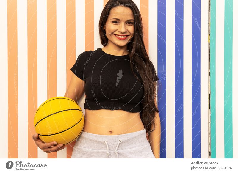 Lächelnde Frau, die mit einem Basketball an einer gestreiften Wand steht Sportlerin Ball Wellness Hobby Freizeit physisch Spieler Motivation sportlich passen