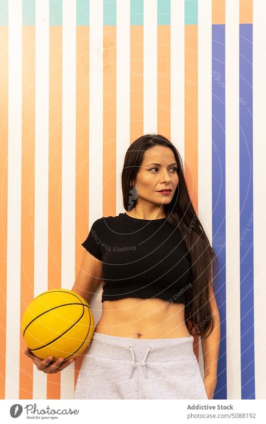 Nachdenkliche Frau, die mit einem Basketball an einer gestreiften Wand steht Sportlerin Ball Wellness Hobby Freizeit physisch Spieler Motivation sportlich