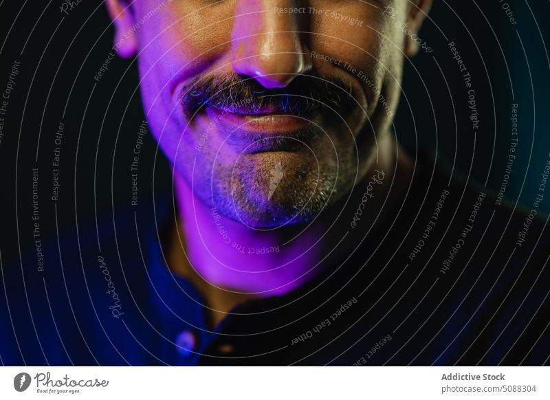 Crop Gesicht eines Mannes mit Schnurrbart gegen schwarzen Hintergrund glühen neonfarbig Licht leuchten maskulin Studioaufnahme Dunkelheit Lächeln positiv froh