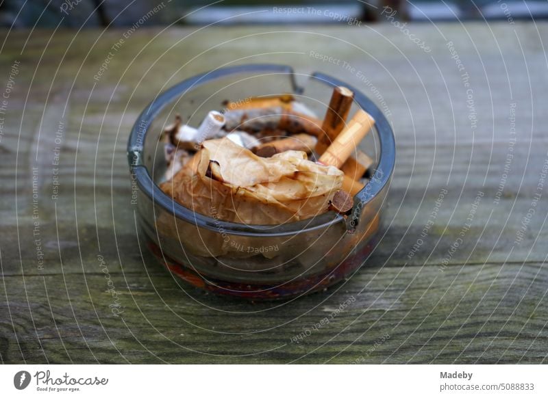 Runder Aschenbecher aus Glas mit Kippen von Zigaretten und buntem Herbstlaub auf einem rustikalen Holztisch mit Maserung im Herbst in den Gassen der Altstadt von Detmold am Teutoburger Wald in Ostwestfalen-Lippe