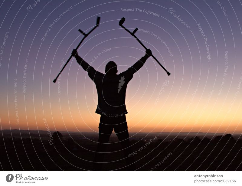 Silhouette eines Mannes, der Krücken hochhält, um das Erreichen der Spitze eines Hügels bei Sonnenuntergang zu feiern geheilt Gesundheit Gesundheitswesen