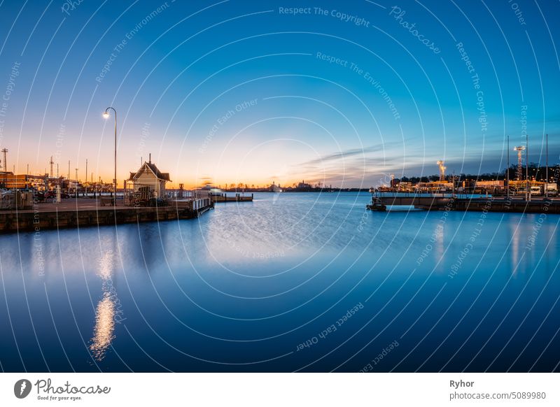 Helsinki, Finnland. Landschaft mit Stadt Pier, Steg bei Sonnenaufgang Zeit. Blauer Himmel reflektiert in ruhigen Meer Wasseroberfläche. Liegeplatz in Beleuchtung am Abend Nachtbeleuchtung