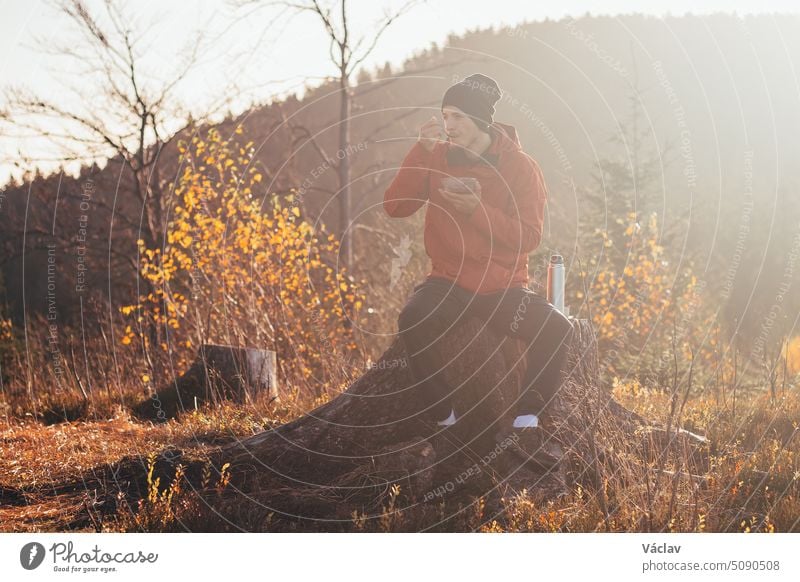 Tourist sitzt auf einem Baumstumpf und isst bei Sonnenaufgang Porridge. Ein Wanderer wird von der Morgensonne erleuchtet und genießt die Aussicht. Beskiden, Tschechische Republik