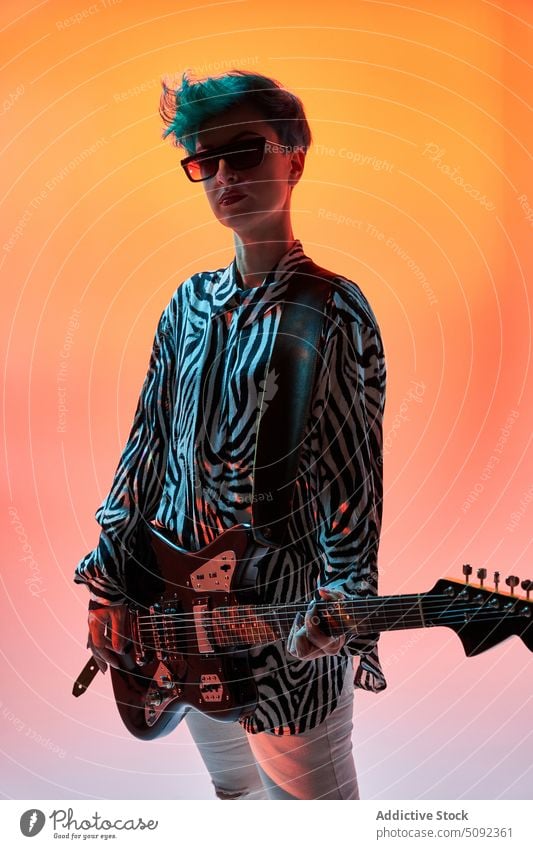 Stilvolle Frau spielt Gitarre in buntem Studio elektrisch spielen Musik Musiker Gitarrenspieler trendy Hipster Mode Outfit Sonnenbrille Instrument Melodie Klang