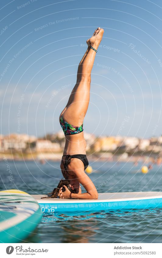 Schlanke Frau in Badekleidung macht Kopfstand auf Paddleboard Zusatzplatine Yoga MEER Wasser ruhig Asana Rippeln aqua schlank üben Gleichgewicht Pose