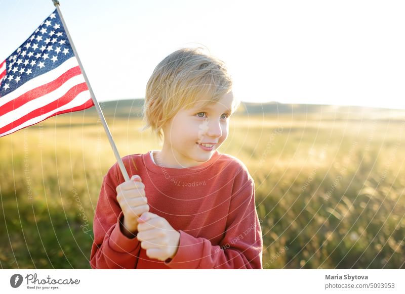 Netter kleiner Junge feiert den 4. Juli, den Unabhängigkeitstag der USA bei sonnigem Sommer Sonnenuntergang. Kind läuft und springt mit amerikanischer Flagge Symbol der Vereinigten Staaten über Weizenfeld. Patriotismus