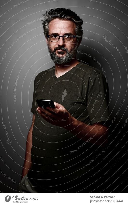 bärtiger Mann mit Brille in T-Shirt mit Smartphone vor einem grauen einfarbigen Hintergrund blickt in die Kamera ernst dunkel Schatten männlich kontrastreich