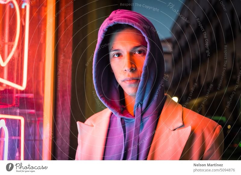 Junger hispanischer Mann der Jahrtausendwende mit rotem Neonlicht und einer Kapuze jung Straße hell Mode Anzug Aussehen Leder Licht Nacht Typ Konzept bunt