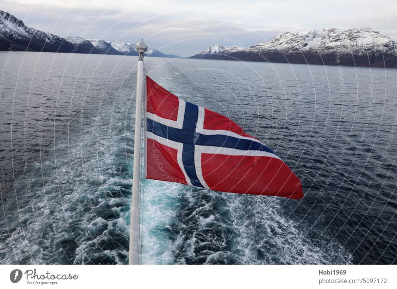 Blick von einem Schiff in einen Fjord bei Alta in Norwegen mit einer Fahne im Vordergrund. Boot Heck Strömung Berg Berge Schnee Wasser Meerwasser Ozean Kalt