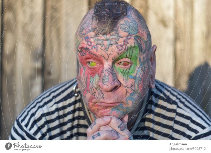 Ein im Gesicht vollständig tätowierter Mann, mit nachdenklichem Gesichtsausdruck. Auch seine Augäpfel sind nicht weiß, denn sie sind mittels Eyeball-Tattoo farbig gestaltet.