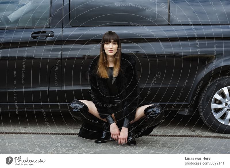Ein wunderschönes brünettes Model posiert für einen Modelltest. Eine modische Frau in Schwarz gekleidet mit einigen Latex-Akzenten und einem schwarzen Auto als Hintergrund. Heiße Reifen und noch heißere Schönheit.