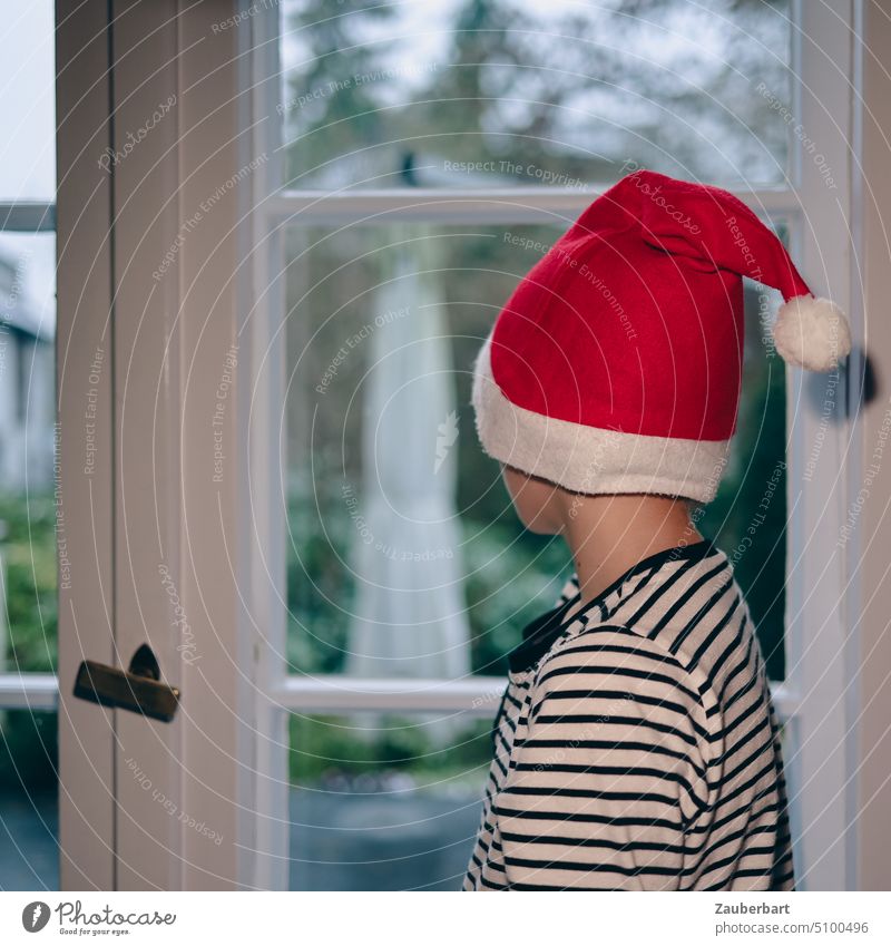 Kleiner Junge mit roter Weihnachts- oder Nikolaus-Mütze im gestreiften Schlafanzug steht am Fenster und wartet auf Weihnachten klein Weihnachtsmann warten weiß