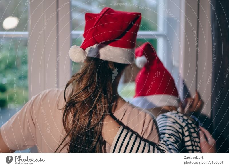 Zwei Kinder mit Weihnachts- oder Nikolaus-Mützen schauen Arm in Arm aus dem Fenster und warten auf Weihnachten Geheimnis Freude Vorfreude rot weiß Kindheit