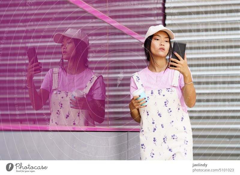 Junge asiatische Frau mit Smartphone und Kaffee in der Nähe von rosa Wand benutzend Straße lesen Nachricht Stil online Gebäude soziale Netzwerke Glaswand