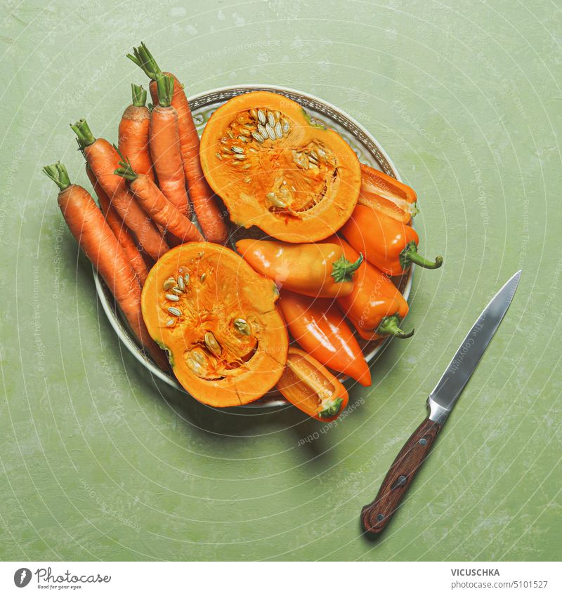 Teller mit orangefarbenem Gemüse: Karotten, Kürbisse, Paprika auf grünem Hintergrund mit Messer, Ansicht von oben. Gesundes Essen, Draufsicht gesunde Ernährung