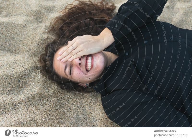 Fröhliche Frau, die ihr Auge bedeckt, während sie im Sand liegt Strand Lügen Freude Küste Freizeit Kälte Auge abdecken Spaß haben heiter Lächeln Glück positiv
