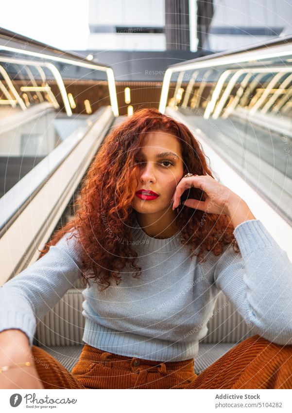 Stilvolle junge Frau sitzt in der Nähe von Rolltreppe Kaufhof Vorschein urban rote Haare krause Haare Gesicht berühren Porträt Rotschopf Pullover Hose Ingwer