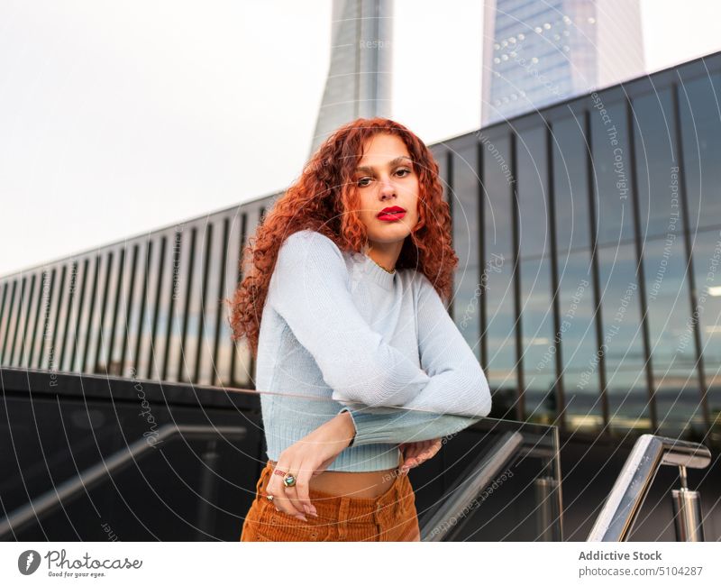 Selbstbewusste rothaarige Frau, die sich an einen Glaszaun auf einer Straße in der Stadt lehnt selbstbewusst Stil Revier Großstadt cool tausendjährig ernst