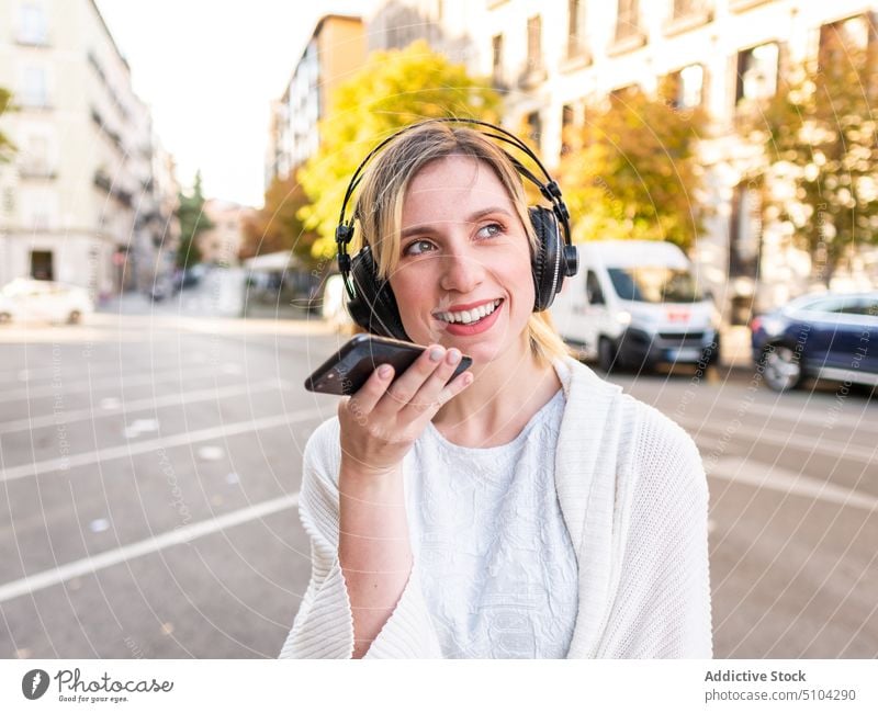 Glückliche junge Frau mit Kopfhörern, die ihr Smartphone benutzt und auf der Straße lächelt Sprachmeldung Aufzeichnen positiv Großstadt Musik Lächeln Audio