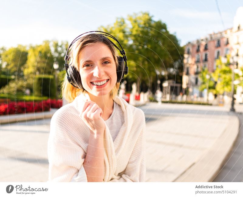 Positiv gestimmte Frau lächelt, während sie auf der Straße mit Kopfhörern ihr Lieblingslied hört Musik zuhören Park Lächeln Großstadt Glück meloman Porträt