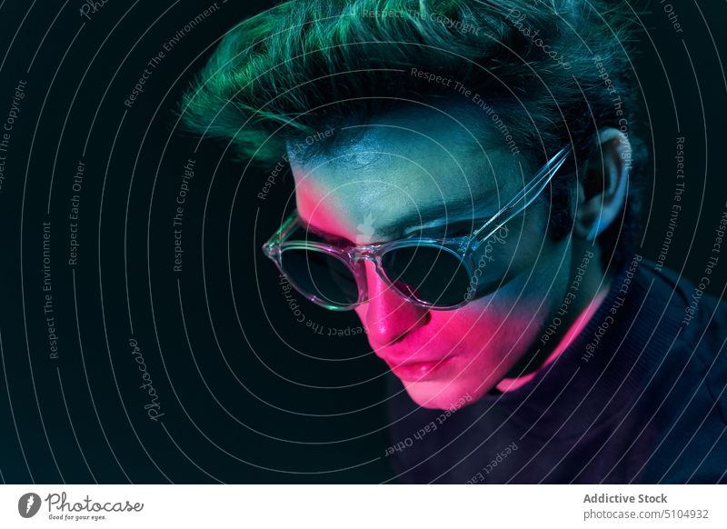 Junges männliches Modell mit Sonnenbrille im Studio stehend Mann neonfarbig Licht Studioaufnahme dunkel Individualität emotionslos Porträt jung leuchtend