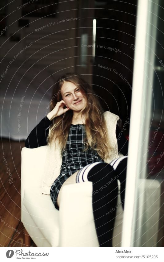 Portrait durch die Balkontür einer jungen blonden Frau, die schräg in einem Sessel sitzt junge Frau Beine Kniestrümpfe Kleid lächeln Freude Gelassenheit zuhause
