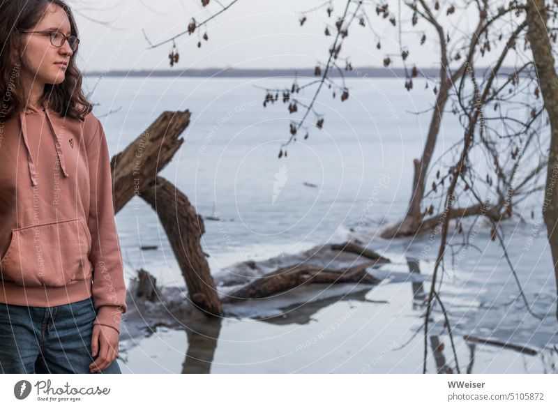 Ein junges Mädchen steht nachdenklich am See, dessen Oberfläche von dünnem Eis überzogen ist junge Frau Wasser Winter kalt eisig frostig melancholisch verträumt