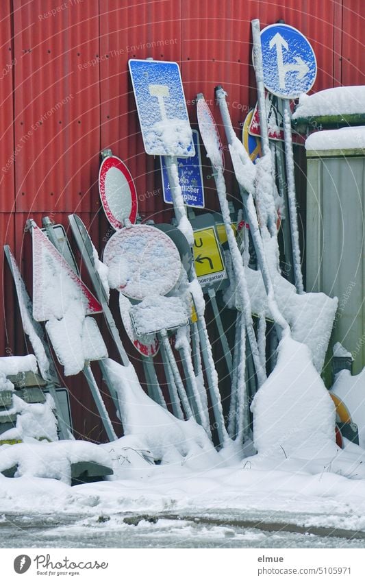 zahlreiche, mit Schnee bedeckte Verkehrszeichen lehnen an einer roten Blechwand / Winter Verkehrsschild Ansammlung Metall StVO Zeichen Schilder