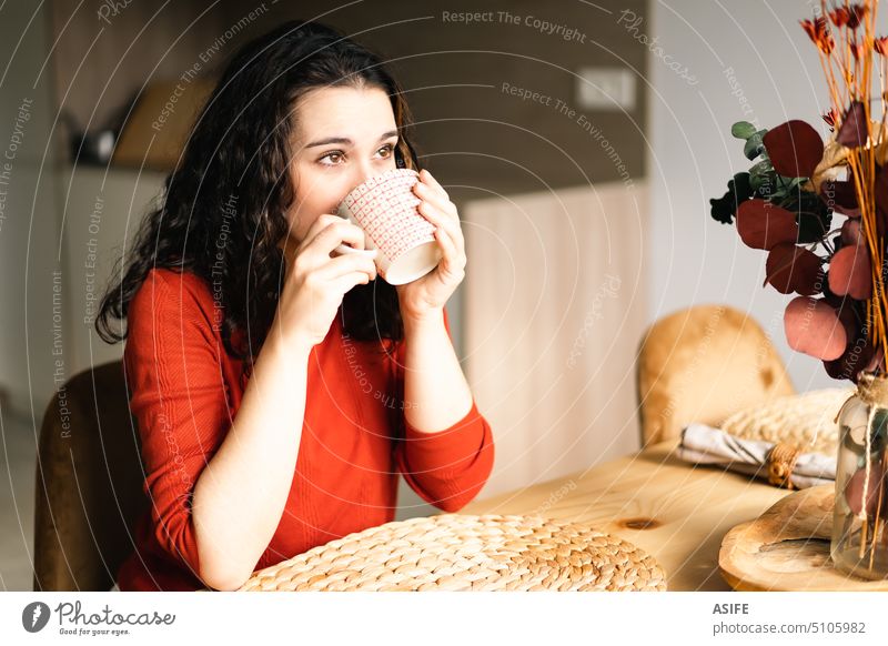 Junge Frau trinkt am Tisch sitzend einen Kaffee Tasse Tee trinken Frühstück Getränk Küche Sitzen Wegsehen verträumt heimwärts Lifestyle Beteiligung heiß Mädchen