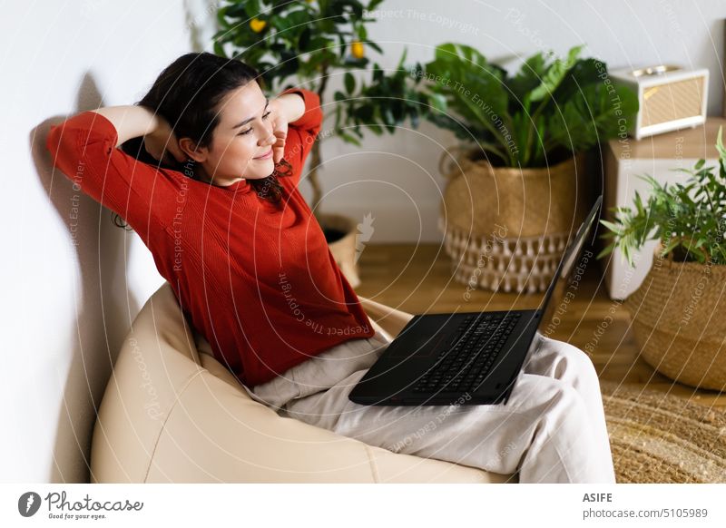 Entspannte junge Frau, die zu Hause in einem Tütenstuhl sitzt und einen Laptop benutzt heimwärts Computer Grasnarbe Liege Tasche Stuhl Glück Freizeit Pflanzen